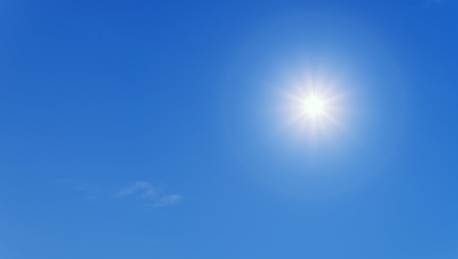 Ein strahlender Sonnenball steht hoch am blauen Himmel, umgeben von wenigen, sanften Wolken. Die Helligkeit der Sonne vermittelt ein Gefühl von Wärme und klarer Tageslichtatmosphäre.