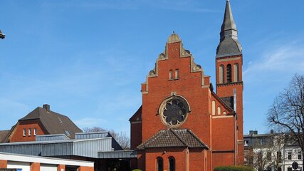 Ein rotes, historisches Kirchengebäude mit einer markanten Spitze und einem großen Rundfenster. Der Himmel ist blau und klar, und die Umgebung ist urban mit weiteren Gebäuden im Hintergrund.