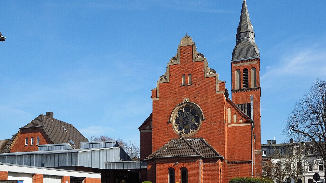 Ein rotes, historisches Kirchengebäude mit einer markanten Spitze und einem großen Rundfenster. Der Himmel ist blau und klar, und die Umgebung ist urban mit weiteren Gebäuden im Hintergrund.