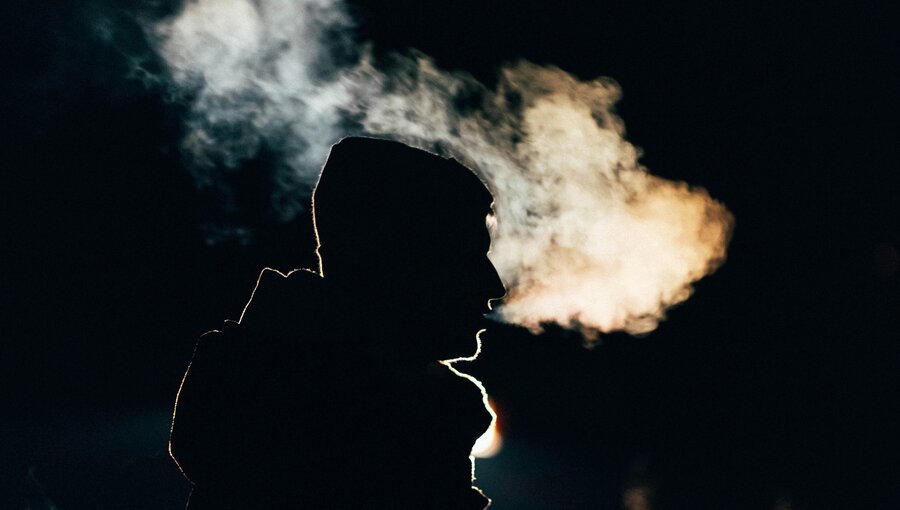 Ein Silhouette einer Person in winterlicher Kleidung steht im Fokus, während Rauch oder Nebel aus ihrem Mund aufsteigt. Der Hintergrund ist dunkel und unscharf, was die Atmosphäre der Nacht verstärkt.
