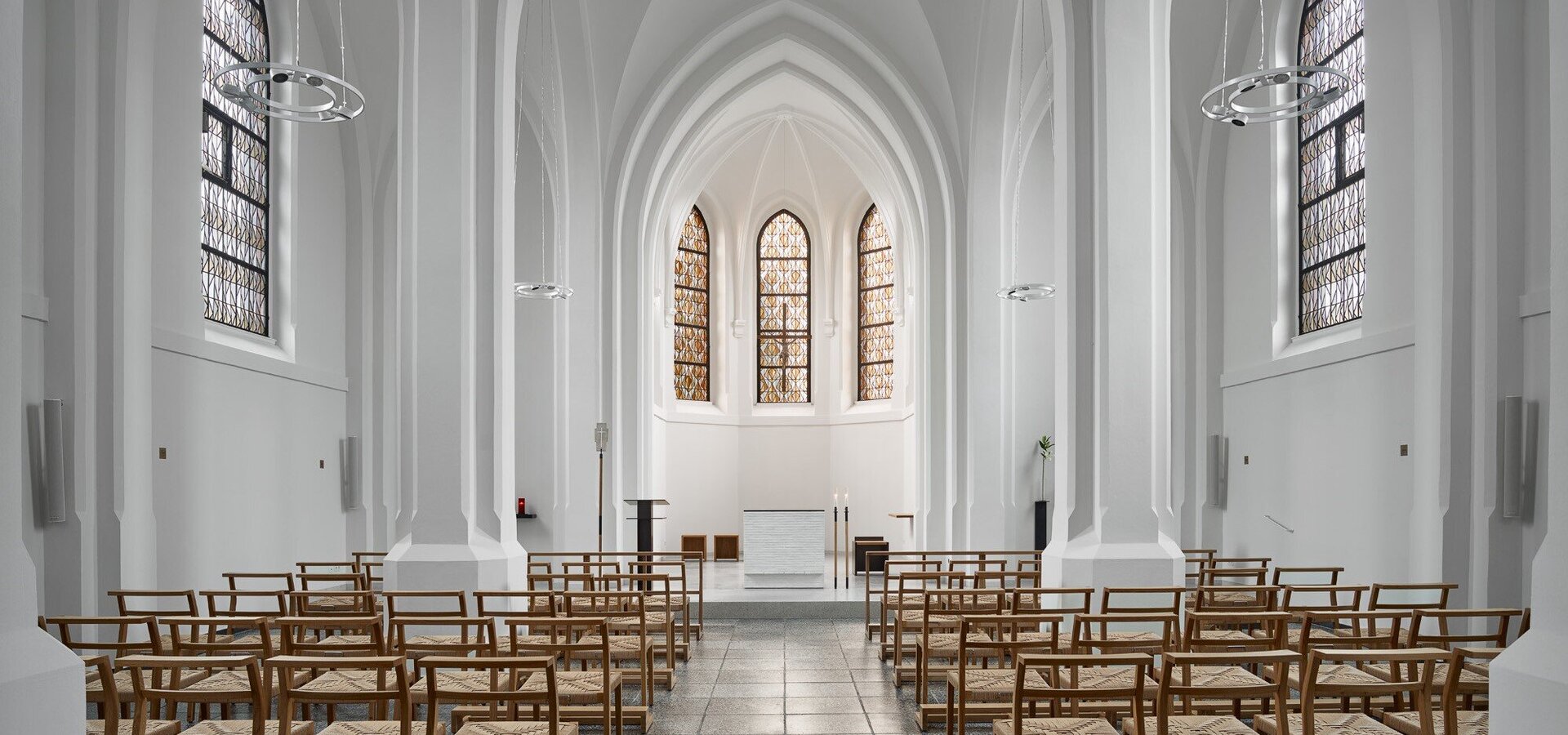 Eine minimalistische Kirche mit hohen, weißen Wänden und großen Fenstern, die ein sanftes Licht hereinlassen. In der Mitte stehen Reihen von Holzstühlen. Altar und Kanzel sind am vorderen Ende sichtbar, was eine ruhige und einladende Atmosphäre schafft.