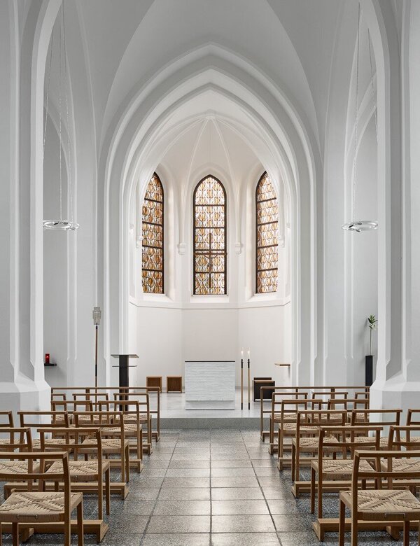 Eine minimalistische Kirche mit hohen, weißen Wänden und großen Fenstern, die ein sanftes Licht hereinlassen. In der Mitte stehen Reihen von Holzstühlen. Altar und Kanzel sind am vorderen Ende sichtbar, was eine ruhige und einladende Atmosphäre schafft.