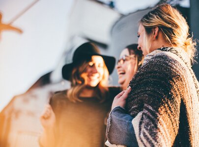 Drei Frauen stehen eng beisammen und lachen miteinander. Eine trägt einen Hut, während die andere eine Brille hat. Sie scheinen sich in einer fröhlichen, entspannten Atmosphäre zu befinden, umgeben von sanftem Licht.