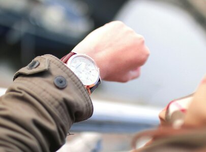 Ein Handgelenk mit einer eleganten Armbanduhr, die auf die Uhrzeit blickt. Die Person trägt eine braune Jacke mit einem Knopf. Im Hintergrund sind unscharfe, natürliche Elemente zu erkennen, die auf eine Außenszene hinweisen.