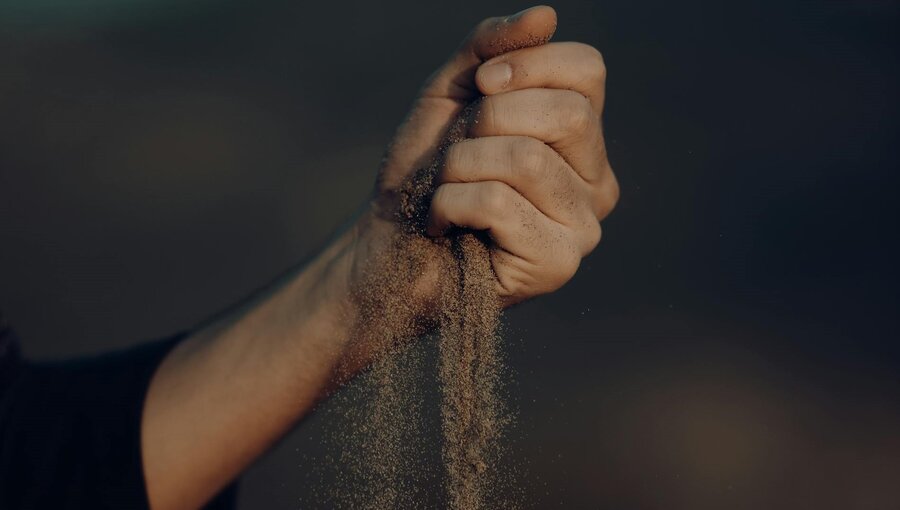 Eine menschliche Hand hält eine Handvoll Sand, der langsam durch die Finger rieselt. Der Fokus liegt auf der Textur des Sands und der Bewegung des Herunterfallens, was ein Gefühl von Vergänglichkeit vermittelt.