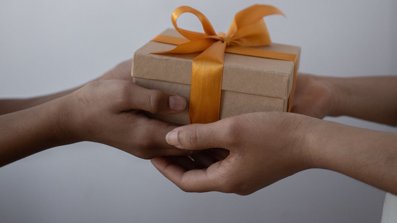Zwei Hände überreichen sich ein Geschenk, das in braunes Papier eingewickelt und mit einem orangefarbenen Band verziert ist. Die Geste vermittelt den Moment des Schenkens und der Freude.