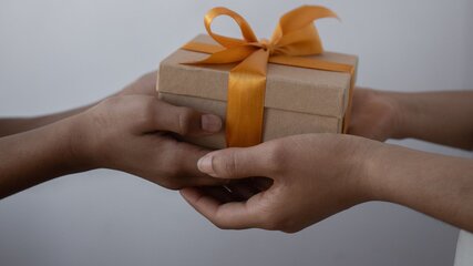 Zwei Hände überreichen sich ein Geschenk, das in braunes Papier eingewickelt und mit einem orangefarbenen Band verziert ist. Die Geste vermittelt den Moment des Schenkens und der Freude.