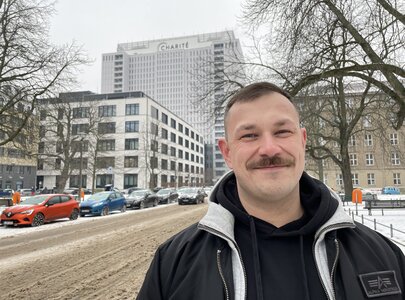 Ein Mann mit einem Schnurrbart steht in einer winterlichen Umgebung auf einer schneebedeckten Straße. Im Hintergrund ist ein modernes Gebäude mit der Aufschrift "Charité" sichtbar. Bäume umrahmen die Szene, und Parkplätze sind links und rechts sichtbar.