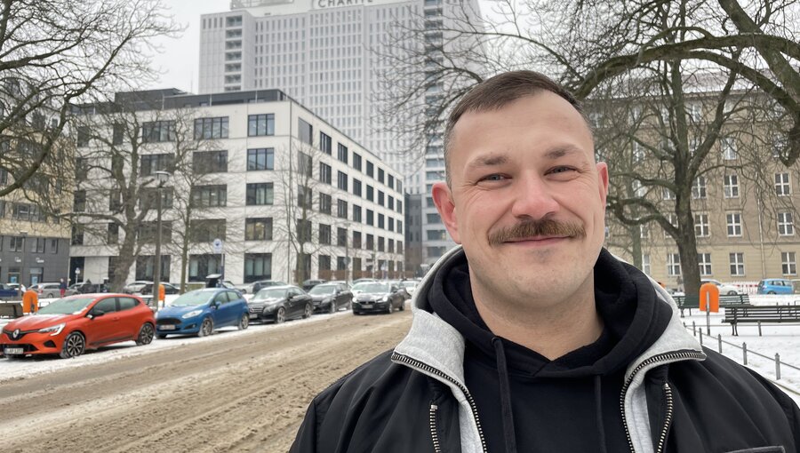 Ein Mann mit einem Schnurrbart steht in einer winterlichen Umgebung auf einer schneebedeckten Straße. Im Hintergrund ist ein modernes Gebäude mit der Aufschrift "Charité" sichtbar. Bäume umrahmen die Szene, und Parkplätze sind links und rechts sichtbar.