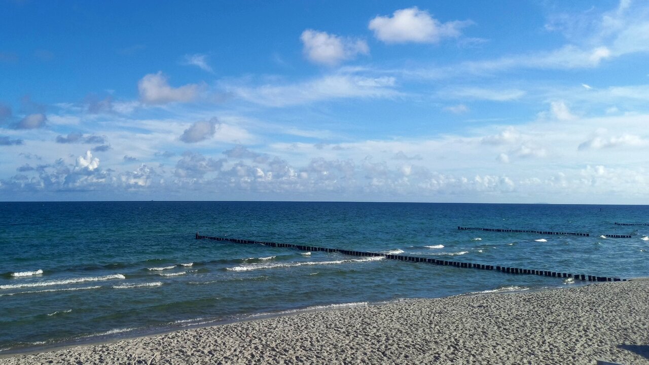 Ein ruhiger Strandszene mit feinem Sand und sanften Wellen. Im Hintergrund erstreckt sich der blaue Ozean unter einem klaren Himmel, der mit einigen weißen Wolken geschmückt ist. Holzpfähle sind parallel zum Ufer im Wasser angeordnet.