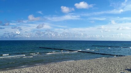 Ein ruhiger Strandszene mit feinem Sand und sanften Wellen. Im Hintergrund erstreckt sich der blaue Ozean unter einem klaren Himmel, der mit einigen weißen Wolken geschmückt ist. Holzpfähle sind parallel zum Ufer im Wasser angeordnet.