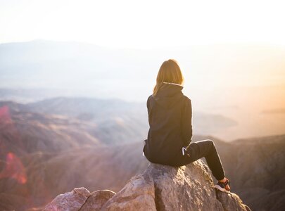 Eine Person sitzt auf einem Felsen und blickt in die Ferne, während die Sonne aufgeht. Die Umgebung besteht aus sanften Hügeln und Bergen, die im warmen Licht der Morgendämmerung beleuchtet werden. Die Szenerie vermittelt ein Gefühl von Frieden und Weite.