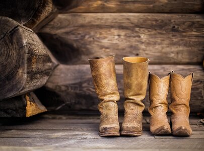 Drei Paar braune Stiefel stehen nebeneinander auf einem Holzfußboden. Die linken Stiefel sind höher und haben ein einfaches Muster, während die rechten etwas kürzer sind und ein detailreicheres Design aufweisen. Im Hintergrund ist die Holzstruktur einer Wand zu sehen.