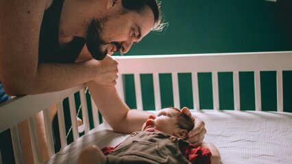 Ein Vater lehnt sich über ein Bettchen, in dem ein Baby liegt. Sie schauen sich liebevoll an, während das Baby entspannt liegt. Das Zimmer hat eine gemütliche Atmosphäre mit einem grünen Farbschema und sanften Lichtquellen.