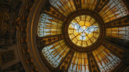 Die Abbildung zeigt ein kunstvolles, rundes Fenster mit einem zentralen Motiv eines Engels. Das Fenster besteht aus vielen bunten, goldenen und gelben Glasstücken, die ein warmes Licht spenden und eine beeindruckende Atmosphäre schaffen.