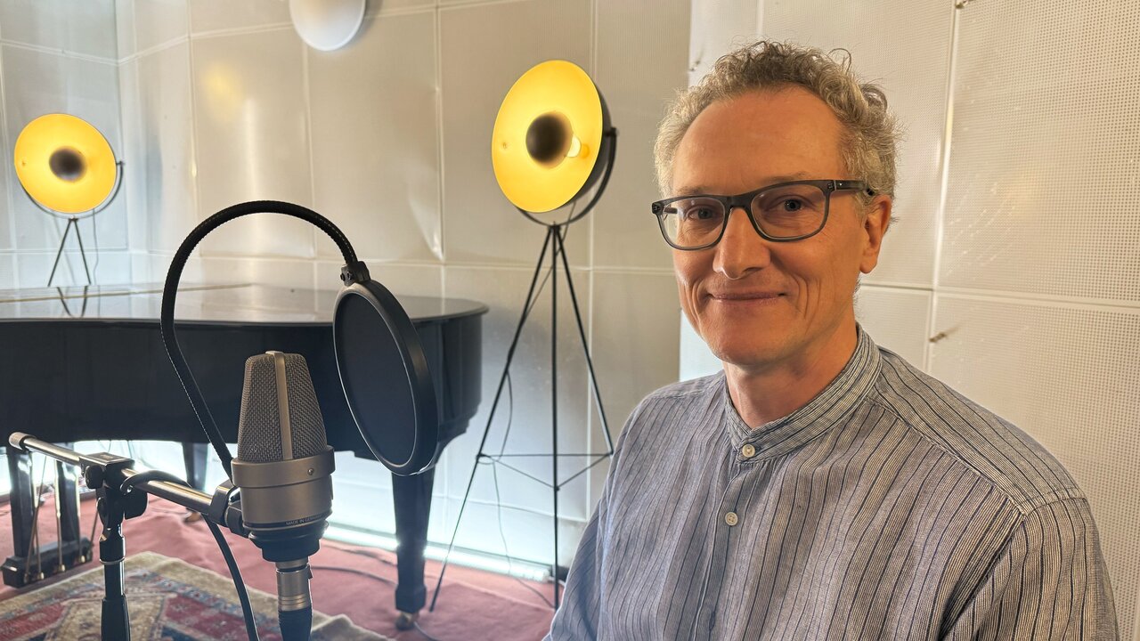 Ein Mann mit Brille sitzt lächelnd vor einem Mikrofon in einem Tonstudio. Im Hintergrund steht ein Piano, und besondere Lampen schaffen eine warme Atmosphäre. Der Mann trägt ein gestreiftes Hemd und wirkt konzentriert und freundlich.
