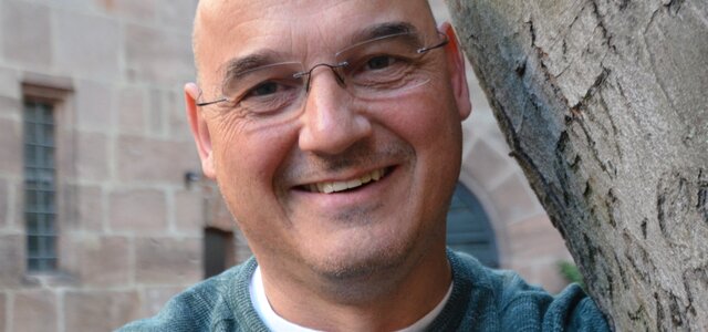 Ein Mann mit Glatze und Brille lächelt freundlich. Er trägt einen grauen Pullover und steht an einem Baumstamm. Der Hintergrund zeigt eine historische Mauer, die das Bild sanft umrahmt.
