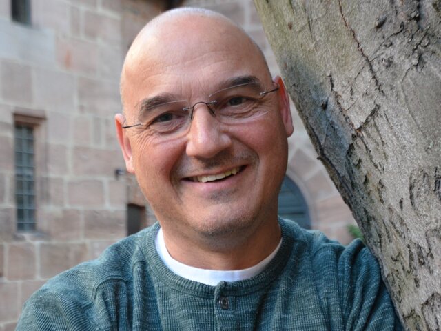 Ein Mann mit Glatze und Brille lächelt freundlich. Er trägt einen grauen Pullover und steht an einem Baumstamm. Der Hintergrund zeigt eine historische Mauer, die das Bild sanft umrahmt.