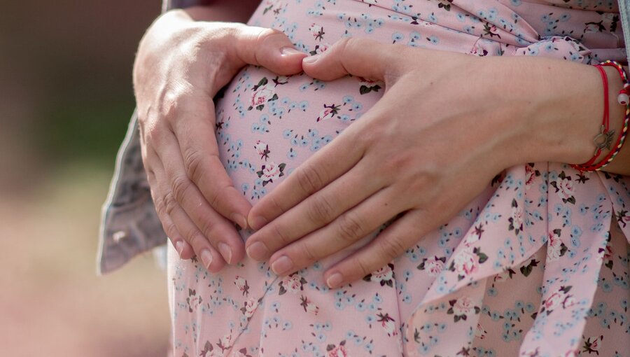 Eine schwangere Person hält mit beiden Händen ihren Bauch, dabei formen die Hände ein Herz. Sie trägt ein blumiges Kleid und die Umgebung ist unscharf, was den Fokus auf die Geste und die Schwangerschaft lenkt.