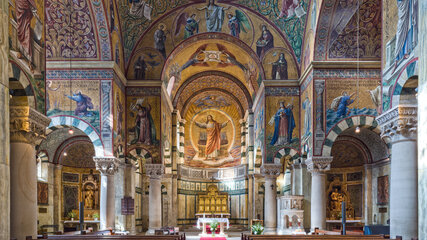 Ein Blick in eine prächtig verzierte Kirche mit hohen Gewölben und bunten Mosaiken. Der Altar ist am Ende des Raumes sichtbar, umgeben von Bänken. Ein roter Teppich führt zum Altar, der den Eindruck von Ehrfurcht und Spiritualität vermittelt.