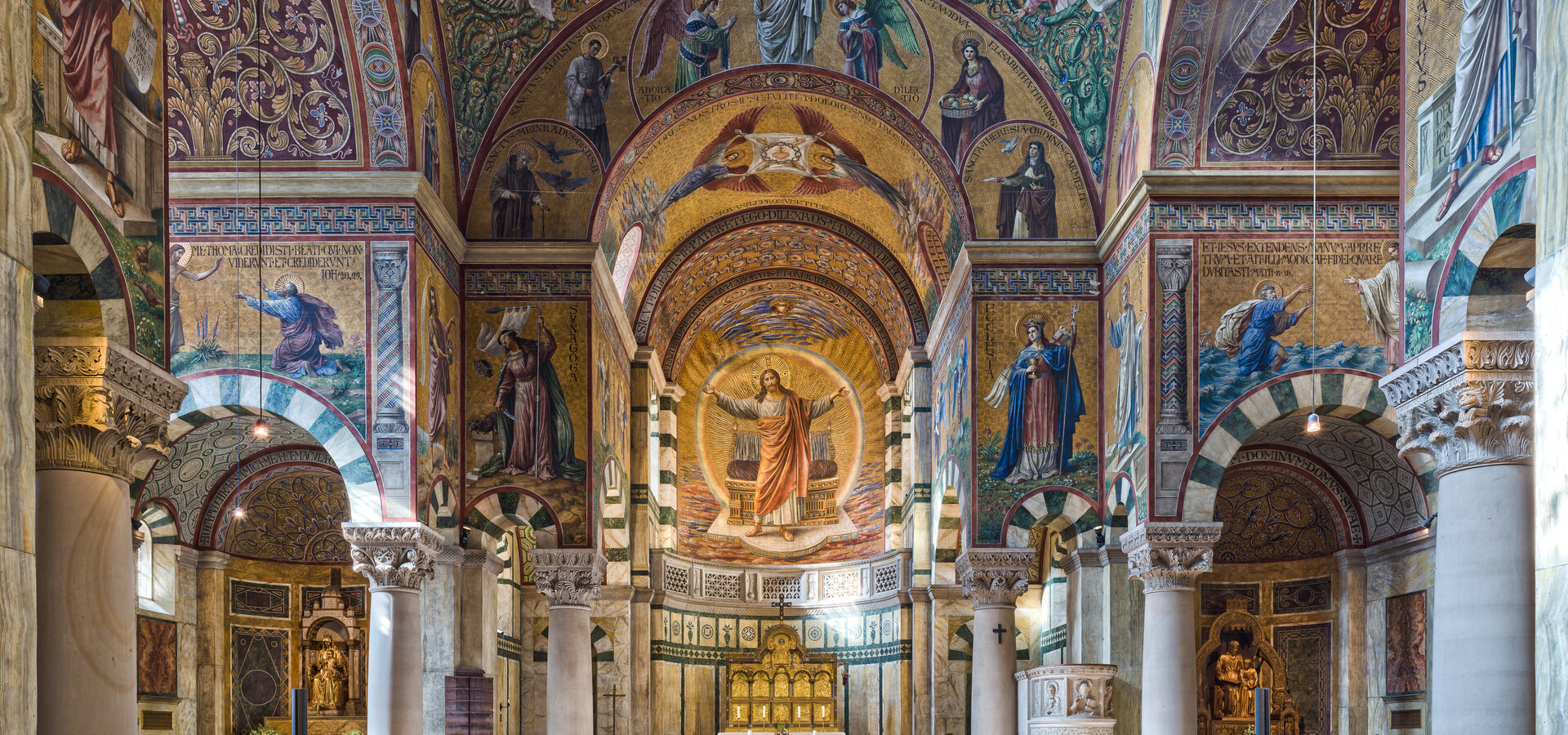 Ein Blick in eine prächtig verzierte Kirche mit hohen Gewölben und bunten Mosaiken. Der Altar ist am Ende des Raumes sichtbar, umgeben von Bänken. Ein roter Teppich führt zum Altar, der den Eindruck von Ehrfurcht und Spiritualität vermittelt.