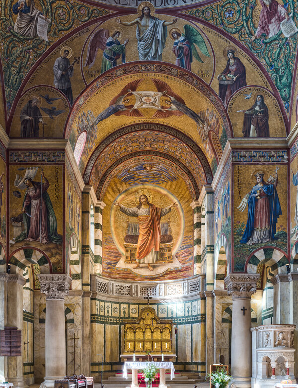 Ein Blick in eine prächtig verzierte Kirche mit hohen Gewölben und bunten Mosaiken. Der Altar ist am Ende des Raumes sichtbar, umgeben von Bänken. Ein roter Teppich führt zum Altar, der den Eindruck von Ehrfurcht und Spiritualität vermittelt.