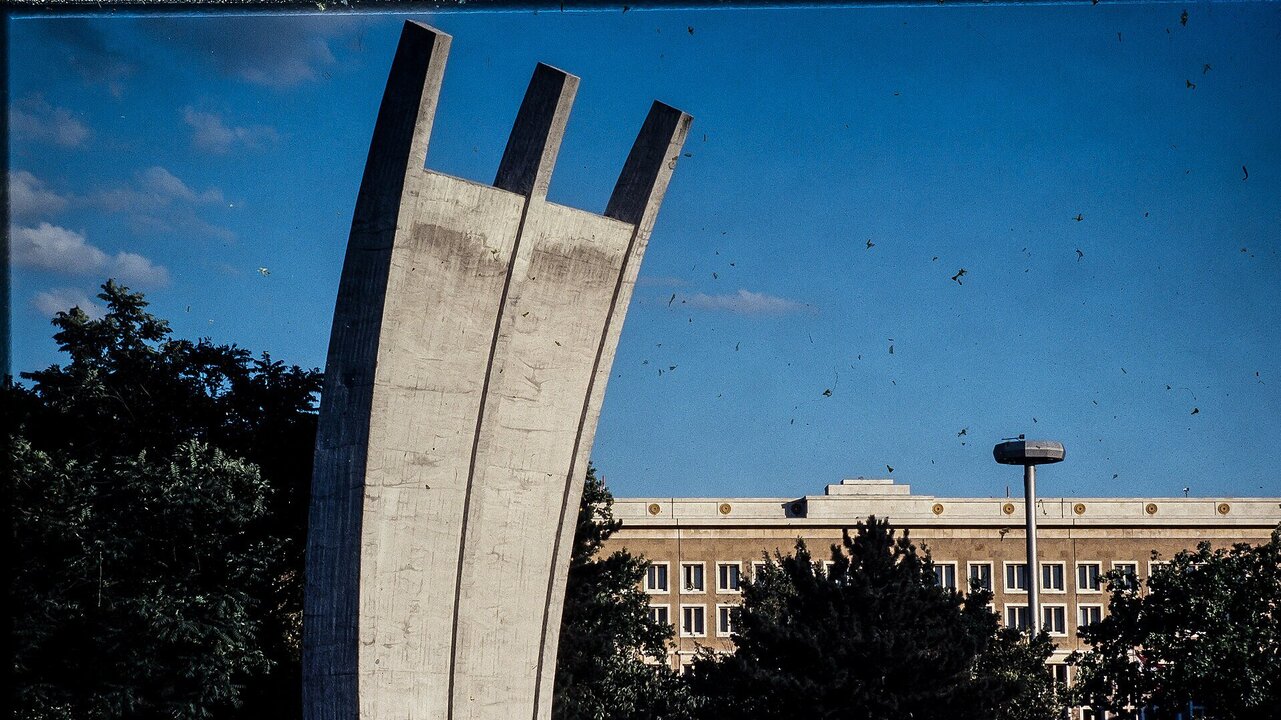Eine große, moderne Betonskulptur, die aus drei nach oben geschwungenen Bögen besteht, steht im Vordergrund. Im Hintergrund ist ein mehrstöckiges Gebäude mit Fenstern und einer Laterne sichtbar, umgeben von Bäumen und Sträuchern bei klarem Himmel.