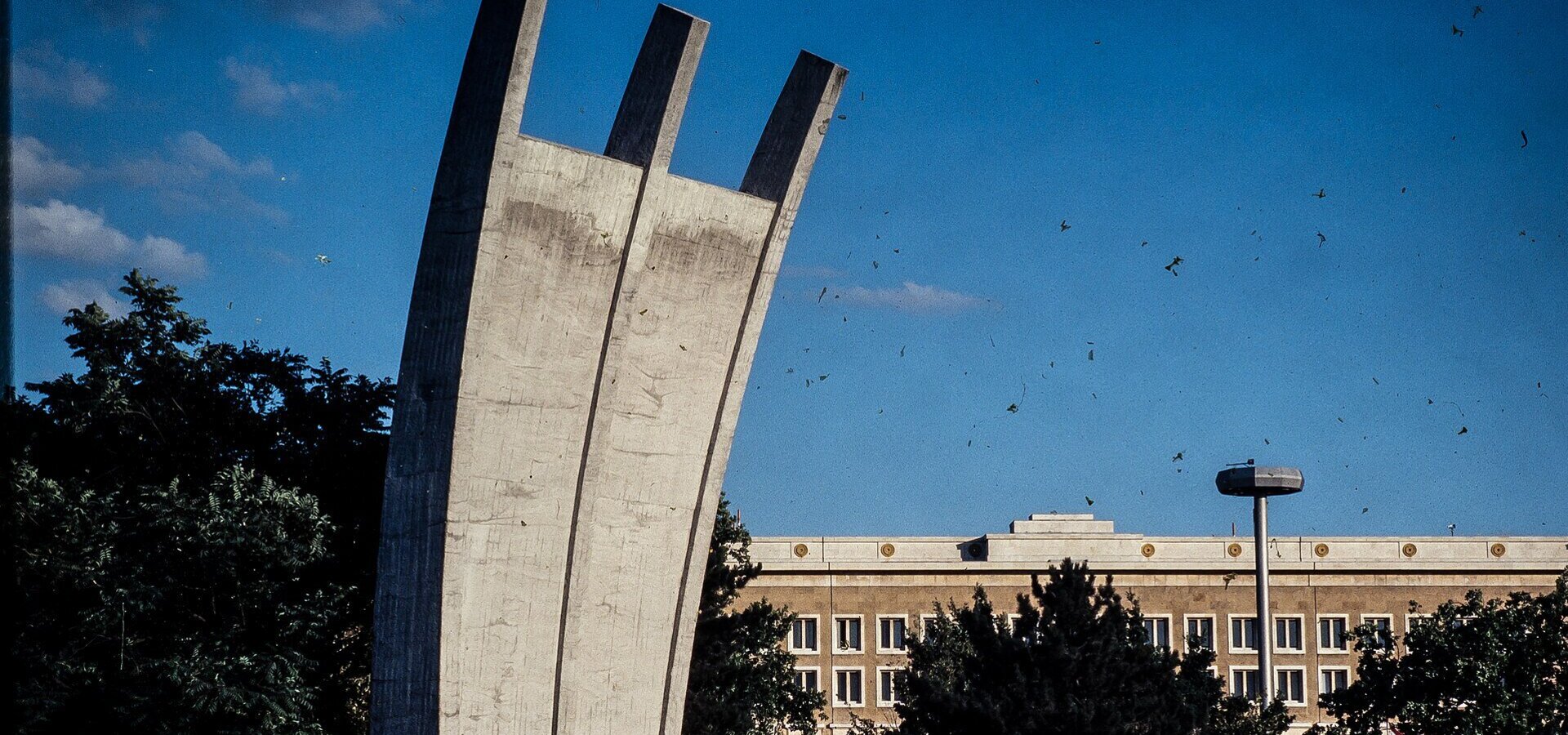 Eine große, moderne Betonskulptur, die aus drei nach oben geschwungenen Bögen besteht, steht im Vordergrund. Im Hintergrund ist ein mehrstöckiges Gebäude mit Fenstern und einer Laterne sichtbar, umgeben von Bäumen und Sträuchern bei klarem Himmel.