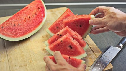 Auf einem Holzbrett liegen mehrere frisch geschnittene Scheiben einer Wassermelone, während eine Hand eine der Scheiben anhebt. Im Hintergrund ist eine frische, halbierte Wassermelone zu sehen, daneben ein Messer.