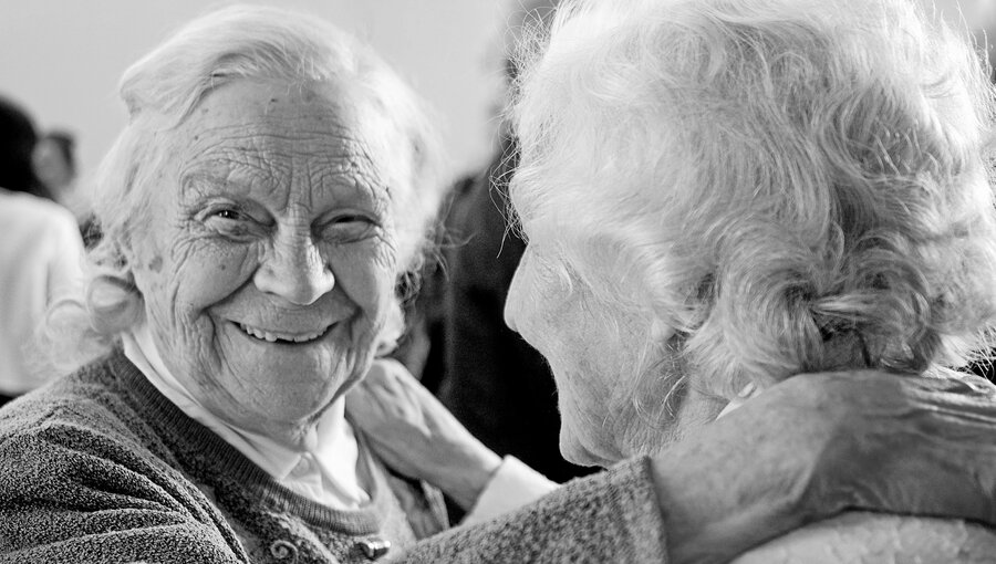 Zwei ältere Frauen umarmen sich herzlich. Die Frau links lächelt mit leuchtenden Augen und hat graue Haare; die Frau rechts ist ihr zugewandt. Die Szene strahlt Freude und Verbundenheit aus.