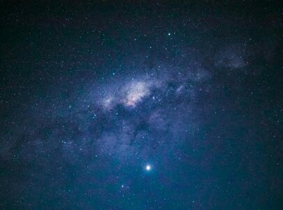 Ein nächtlicher Himmel voll von Sternen. In der Mitte ist eine helle, spiralförmige Galaxie mit einem sanften Farbübergang zwischen Blau und Violett zu sehen. Hell leuchtende Sterne funkeln um die Galaxie herum, während der Hintergrund in dunklem Blau gehalten ist.
