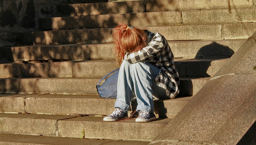 Eine Person mit rotem Haar sitzt traurig auf Steinstufen und umarmt ihre Knie. Die Umgebung zeigt Schatten von Blättern und eine alte Mauer im Hintergrund. Es vermittelt ein Gefühl von Einsamkeit und Nachdenklichkeit.
