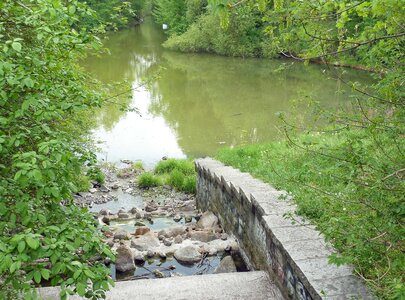 Ein schmaler Weg führt zu einem ruhigen Gewässer, umgeben von dichtem Grün. Steingemauerte Stufen führen hinunter zu einem kleinen Bach, der ins Wasser fließt. Die Szenerie vermittelt eine friedliche, naturnahe Atmosphäre.