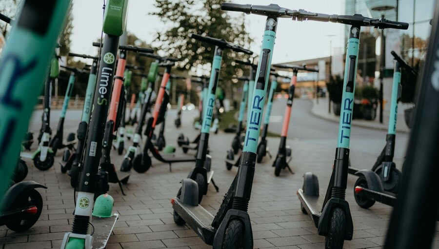 Eine Ansammlung von E-Scootern steht auf einem Gehweg. Die Scooter sind teils in verschiedenen Farben, darunter grün und orange, und verschiedene Marken sind sichtbar. Die Szene vermittelt ein urbanes Umfeld, das für den Transport in der Stadt genutzt wird.