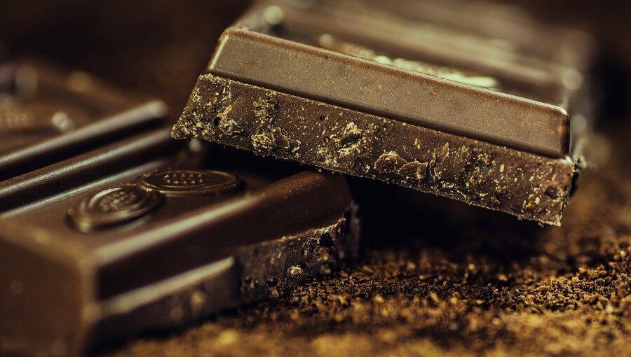 Nahaufnahme von zwei Stückchen dunkler Schokolade, die teilweise übereinander liegen. Die Schokolade weist eine glatte Oberfläche und kleine Krümel auf, die auf eine frische, hochwertige Konsistenz hinweisen. Der Hintergrund ist unscharf und hebt die Schokolade hervor.