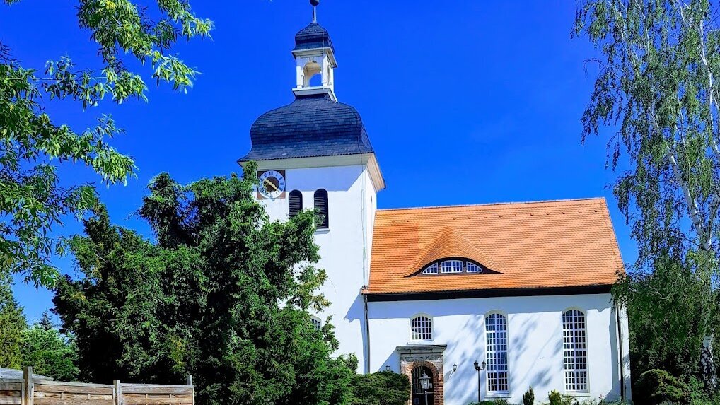 Eine helle Kirche mit einem hohen, schwarzen Turm und einem orangefarbenen Dach steht inmitten von Bäumen und gepflegtem Grün. Ein Gehweg führt zur Eingangstür, die von Büschen flankiert wird. Die Szene vermittelt eine friedliche, ländliche Atmosphäre.