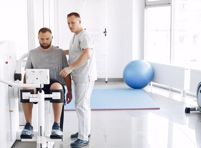Ein Mann sitzt an einem Trainingsgerät, während ein Trainer ihm Anweisungen gibt. Der Raum ist hell und modern, mit Sportgeräten und einer Trainingsmatte im Hintergrund.