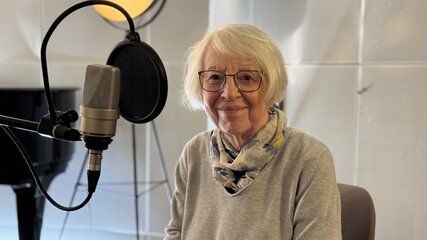 Eine ältere Frau mit Brille und einem Schal sitzt am Tisch in einem Tonstudio. Hinter ihr steht ein Mikrofon, und sie lächelt freundlich in die Kamera. Im Hintergrund ist ein Klavier und eine Beleuchtung sichtbar.