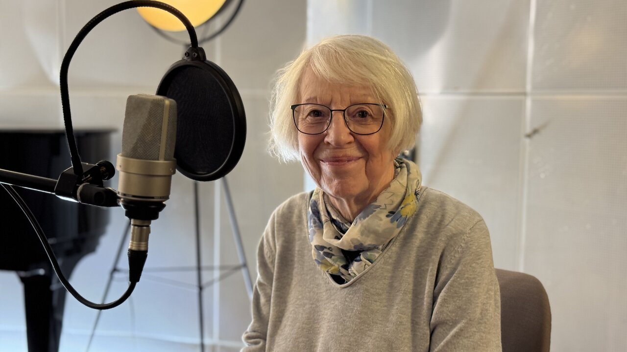 Eine ältere Frau mit Brille und einem Schal sitzt am Tisch in einem Tonstudio. Hinter ihr steht ein Mikrofon, und sie lächelt freundlich in die Kamera. Im Hintergrund ist ein Klavier und eine Beleuchtung sichtbar.