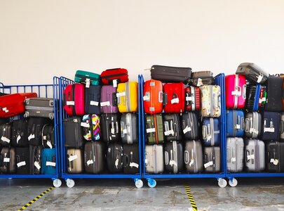 Mehrere Gepäckwagen stehen nebeneinander, gefüllt mit verschiedenen Koffern in unterschiedlichen Farben und Größen. Die Koffer sind gestapelt und mit Gepäckanhängern versehen, was auf eine Gepäckaufbewahrung oder einen Flughafenbereich hinweist.