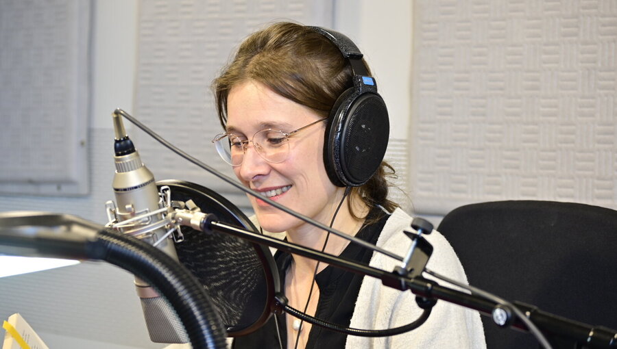 Eine junge Frau mit Kopfhörern lächelt, während sie in ein Mikrofon spricht. Sie hält ein Buch in der Hand und sitzt in einem Studio mit akustischer Dämmung. Ihr Gesichtsausdruck zeigt Konzentration und Freude beim Vorlesen.