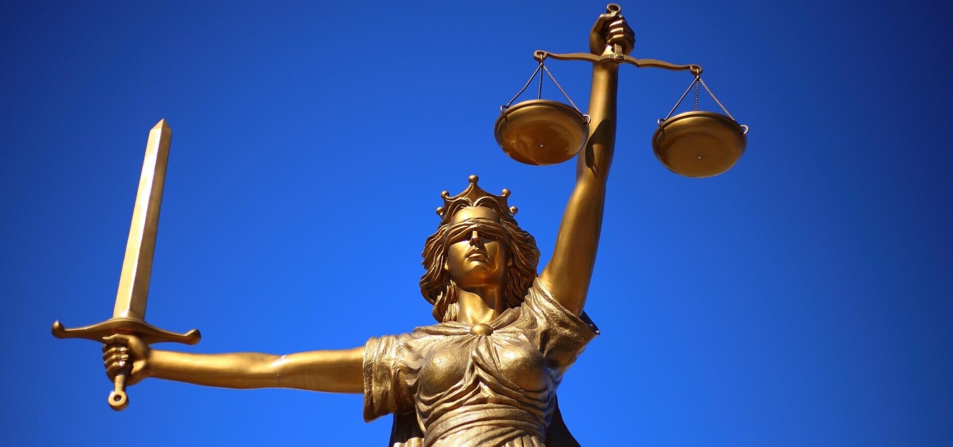 Eine goldene Statue der Justitia steht vor einem blauen Himmel. Sie hält in der rechten Hand eine Waage und in der linken Hand ein Schwert. Die Statue symbolisiert Gerechtigkeit und Gleichgewicht.