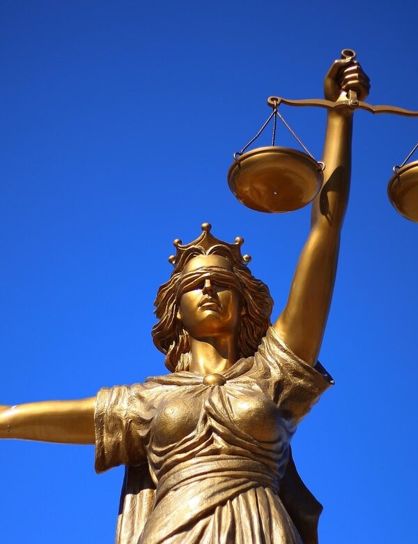 Eine goldene Statue der Justitia steht vor einem blauen Himmel. Sie hält in der rechten Hand eine Waage und in der linken Hand ein Schwert. Die Statue symbolisiert Gerechtigkeit und Gleichgewicht.