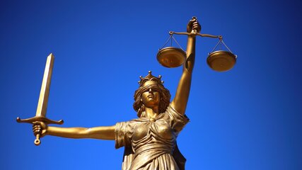 Eine goldene Statue der Justitia steht vor einem blauen Himmel. Sie hält in der rechten Hand eine Waage und in der linken Hand ein Schwert. Die Statue symbolisiert Gerechtigkeit und Gleichgewicht.