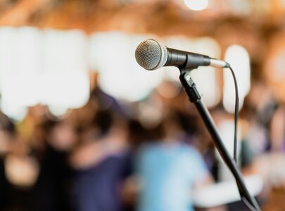 Ein Mikrofon steht im Vordergrund, während im Hintergrund verschwommen eine Gruppe von Menschen zu sehen ist. Die Szene vermittelt eine Atmosphäre eines Interviews oder einer Ansprache in einem Raum voller Teilnehmer.