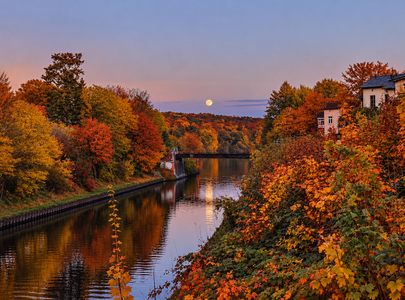 Ein ruhiger Fluss spiegelt die warmen Farben des Herbstes wider. Am Ufer stehen Bäume mit leuchtend orange- und rottönen Blättern. Im Hintergrund ist der Aufgang des Mondes am klaren Abendhimmel zu sehen, während die sanften Hügel den Horizont bilden.