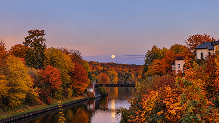 Ein ruhiger Fluss spiegelt die warmen Farben des Herbstes wider. Am Ufer stehen Bäume mit leuchtend orange- und rottönen Blättern. Im Hintergrund ist der Aufgang des Mondes am klaren Abendhimmel zu sehen, während die sanften Hügel den Horizont bilden.