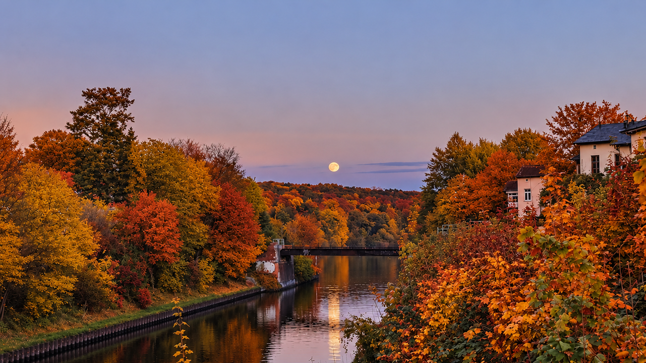 Ein ruhiger Fluss spiegelt die warmen Farben des Herbstes wider. Am Ufer stehen Bäume mit leuchtend orange- und rottönen Blättern. Im Hintergrund ist der Aufgang des Mondes am klaren Abendhimmel zu sehen, während die sanften Hügel den Horizont bilden.
