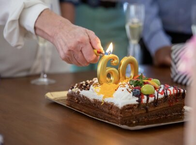 Eine Hand zündet eine Kerze mit der Zahl 60 auf einem Schokoladenkuchen, der mit Sahne, bunten Früchten und Schokoladensplittern dekoriert ist. Der Kuchen steht auf einem Tisch, umgeben von Geschenken und Gläsern mit Sekt.