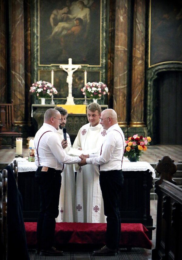 Zwei Männer stehen sich in einer Kirche gegenüber und halten sich an den Händen. Ein Priester, der vor ihnen steht, leitet das Zeremoniell. Im Hintergrund sind Blumen und Kerzen sichtbar, die eine festliche Atmosphäre schaffen.